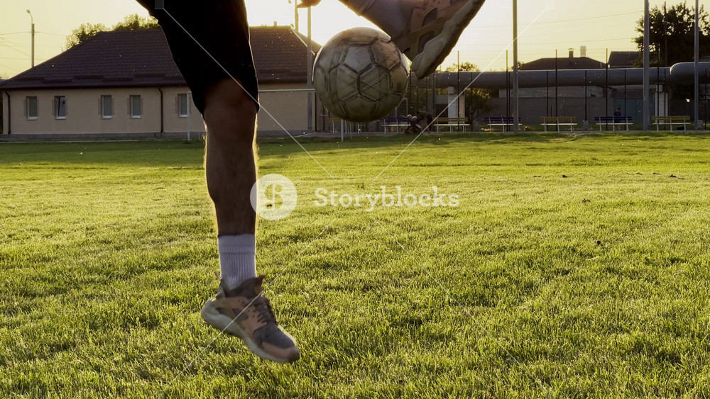 Male feet of professional footballer kicking ball at green field. Legs of young man juggling soccer ball on stadium at sunset. Sportsman