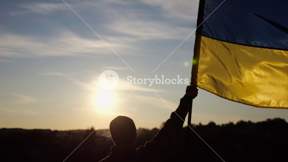 Male ukrainian army soldier lifting blue-yellow banner in honor of the victory against russian aggression at sunset. Young man in camouflage