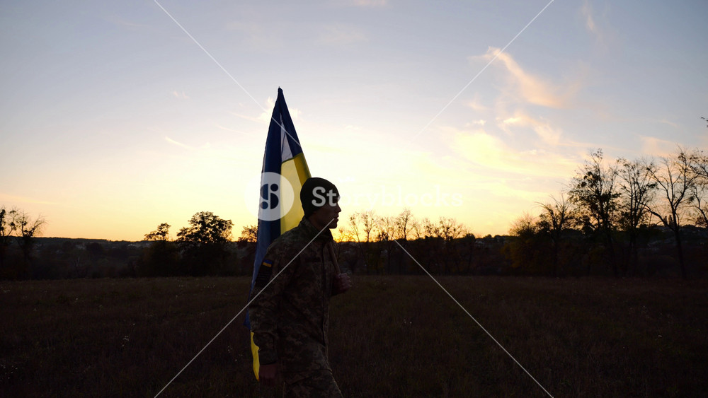 Young male military in uniform walks with flag of Ukraine at meadow. Soldier of ukrainian army going with blue-yellow banner on field at