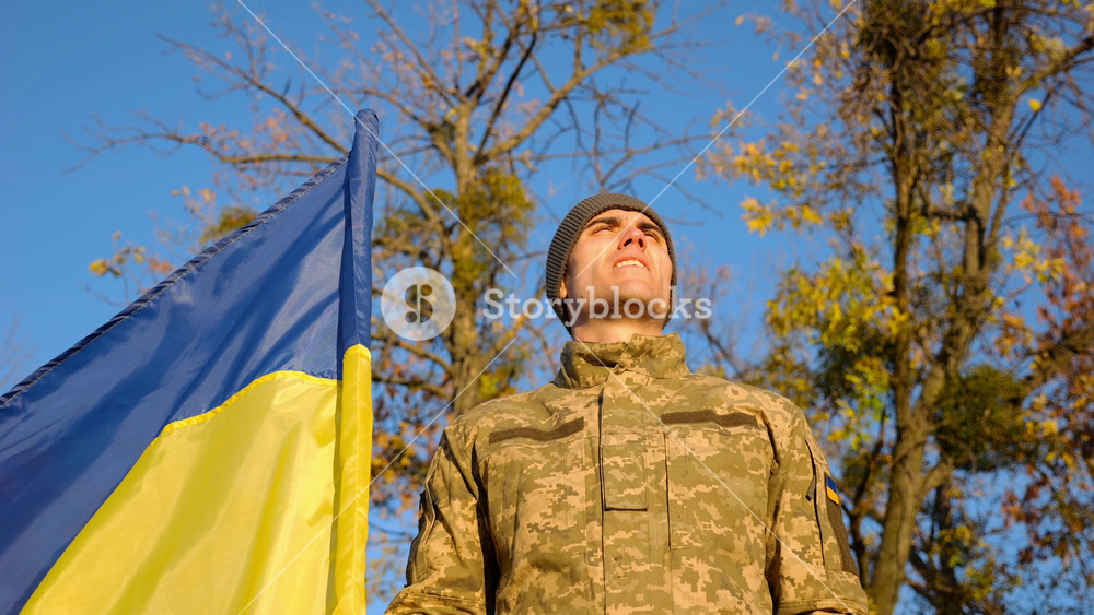 Young man in camouflage uniform stands with flag of Ukraine at countryside against blue sky. Male ukrainian army soldier raising up hand in honor of the victory against russian aggression. Slow motion
