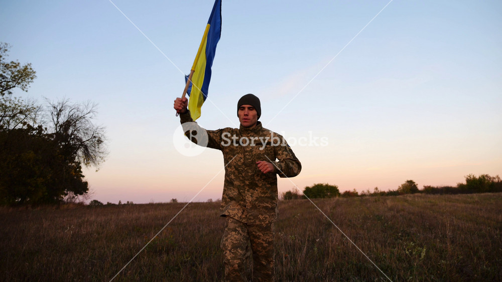 Soldier of ukrainian army running with raised blue-yellow banner on field at dusk. Young male military in uniform jogging with flag of