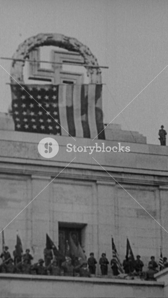 1945 - An American flag is put up over a large swastika symbol at the German Stadium in Nuremberg, Germany, while American soldiers blow up other swastika symbols on the rally grounds.