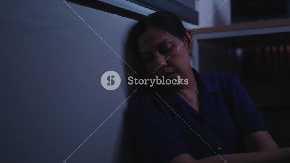 Overwhelmed Woman Leaning on Wall: A woman, appearing weary and burdened, leans her head against a wall in dim lighting, conveying a sense of exhaustion and emotional strain.
