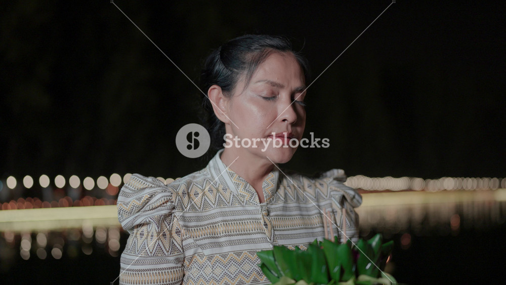 A woman dressed in traditional Thai clothing holds a glowing krathong, her silent prayers reflecting the cultural heritage and spiritual significance of Loy Krathong.