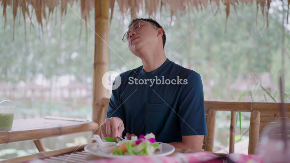 An Asian man holding his belly in pain while eating a meal at a restaurant. He is suffering from a sudden stomachache, indigestion or food poisoning, feeling unwell and unhappy with the bad food.