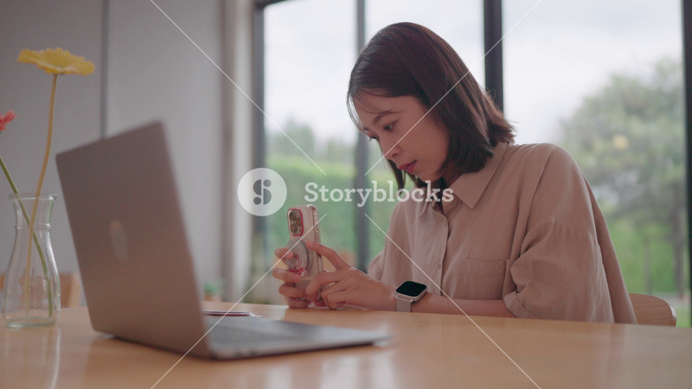 Young woman focused on her phone and laptop. Asian woman is focused on her phone and looking down at the laptop placed in front of her on a wooden table, reminiscent of teleworking or digital communication.