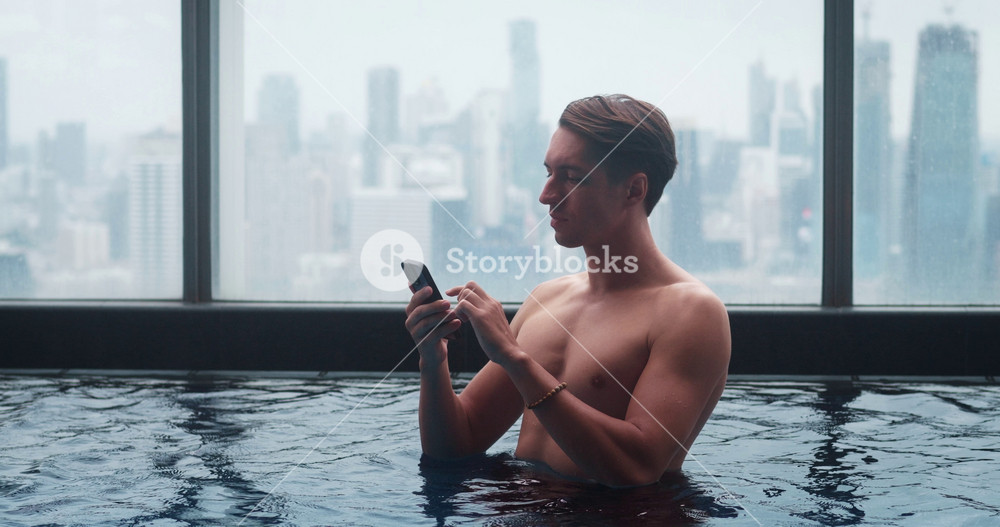 Rich man scrolling mobile phone while relax in roof top swimming pool of in big city. Millennial male tourist using mobile phone in roof top