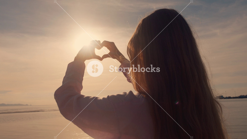 Heart shaped female hands emoji on sunset background. Happy woman made shape of heart enjoys the holiday on the sea. Happiness of freedom on