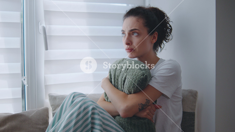 Emotional moment of a woman seated in a cozy living room, embracing a pillow while gazing thoughtfully out the window and looks very lonely