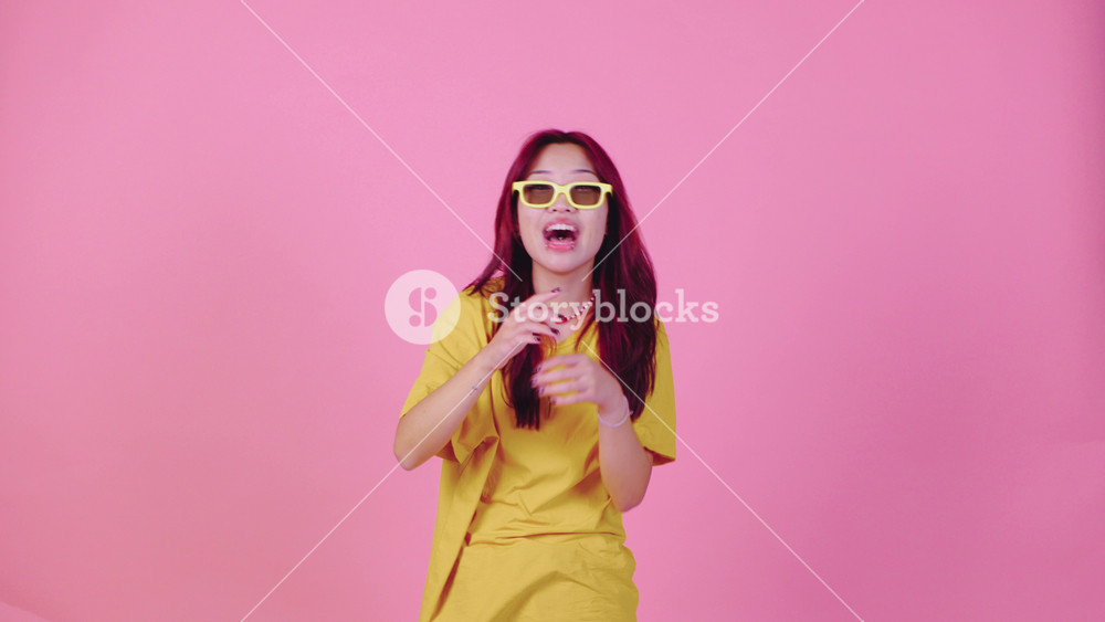 A young Asian woman with vibrant red hair, wearing 3D glasses and a yellow t-shirt, laughs joyfully against a pink background. Her amused expression suggests she s watching a funny movie