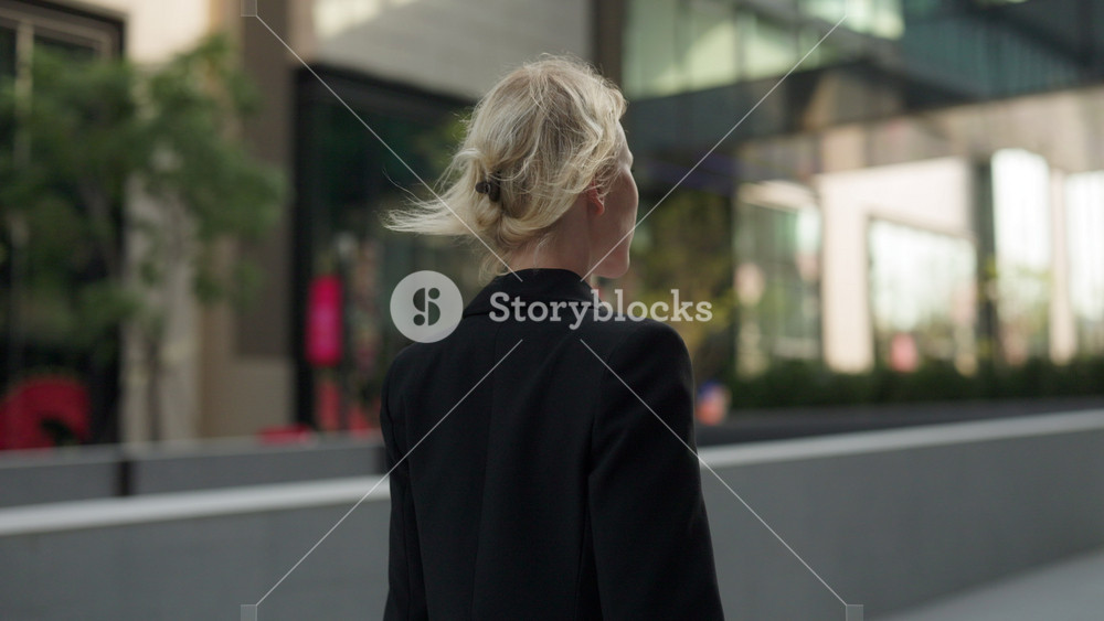 Confident businesswoman in black jacket walks along downtown street backside view. Lady leader with blonde hair strolls alone in shady urban