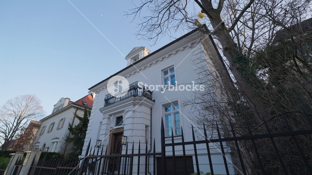 Elegant white building with terrace over entrance near bare trees low angle shot. Old residential house framed by forged metal fence in town
