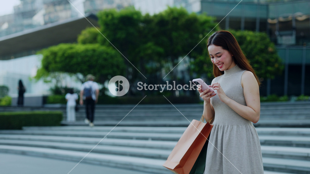 Portrait of Asian woman with shopping bags uses smartphone and smiles in downtown. Pretty Chinese female posts on social media as carries