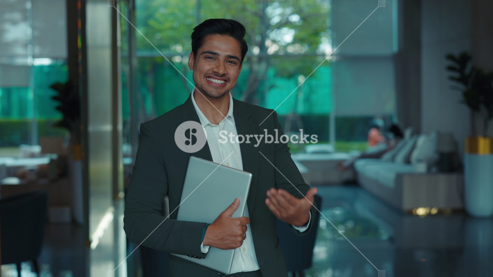 Portrait of positive young Indian businessman with laptop invites customers to corporate co-working premise. Friendly HR manager engages