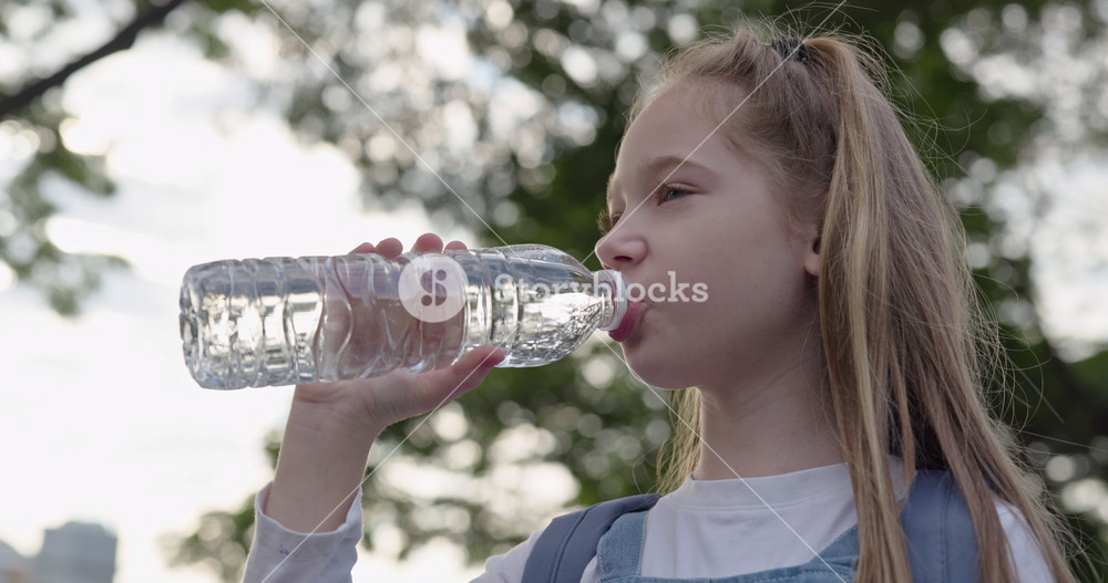 Portrait of cute junior schoolgirl with pigtails drinks water from bottle in park. Little girl with schoolbag refreshes after outdoor