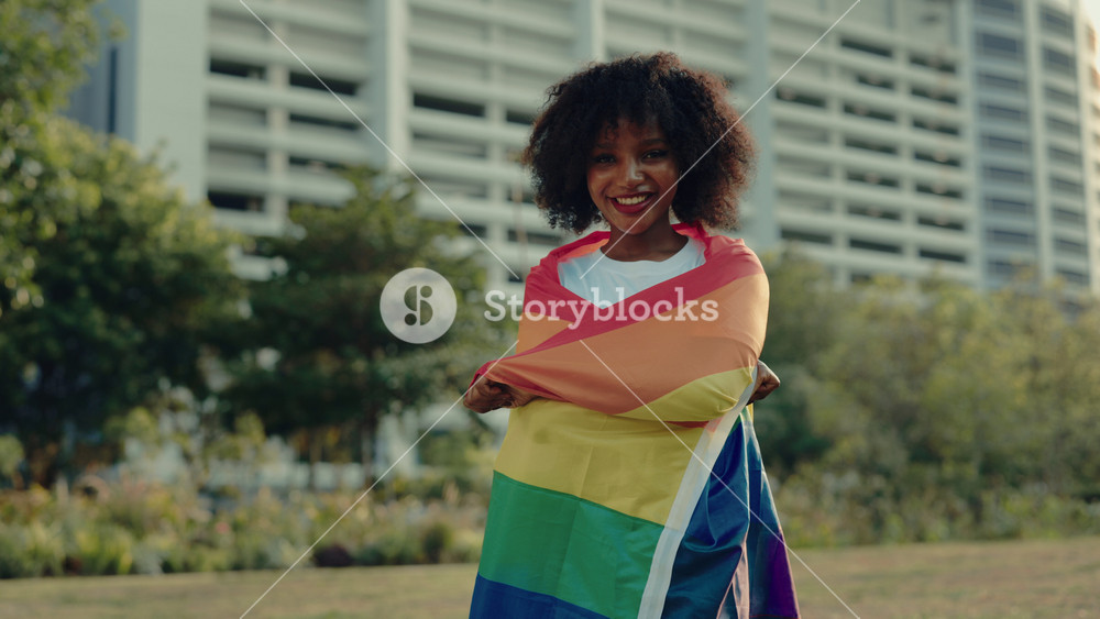 Portrait of smiling freedom woman wrapping with rainbow flag standing near commercial building. Positive African American female proud of