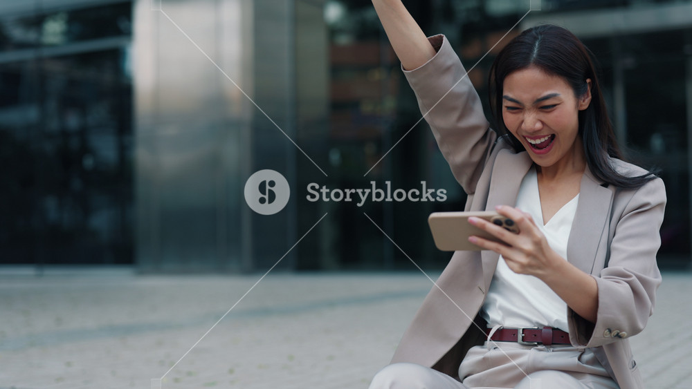 Portrait of Thai woman showing victory gestures while watching live stream on city street. Cheerful Asian woman celebrates success of sports