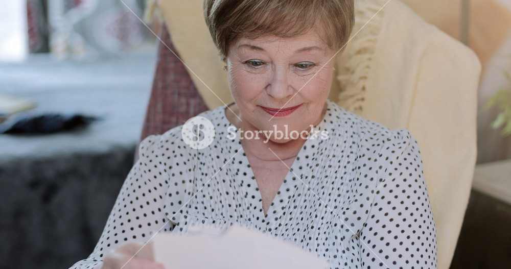 Close up of cheerful grandmother looking at family photos while sitting on chair. Emotional good looking retired female holding old