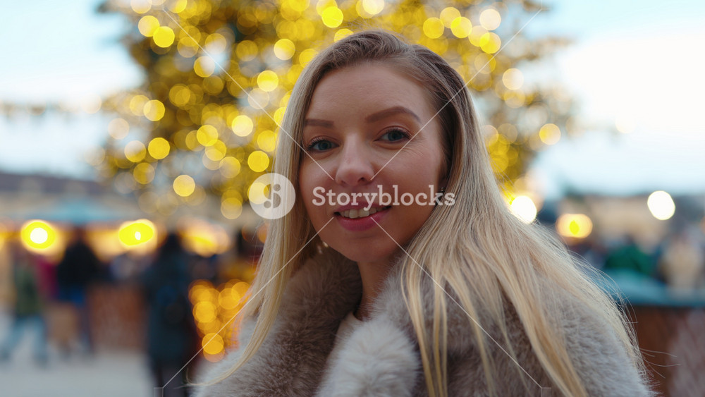 Portrait of smiling woman wearing fur coat stands against booth and Christmas tree. Pretty lady with long hair expresses joy of winter