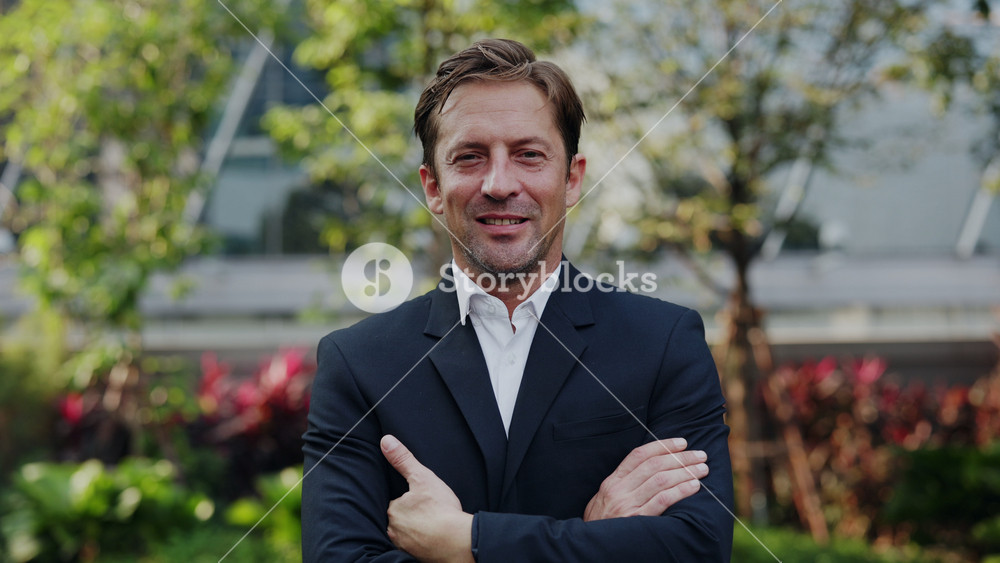 Portrait of happy businessman in suit crosses arms standing in urban park. Mature man entrepreneur smiles conveying success and