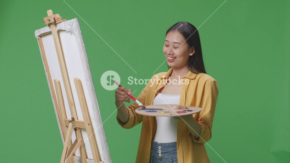 Asian female artist crossing her arms and looking at picture while painting on canvas by oil paints and brush in the green screen studio