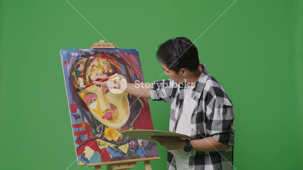 Side view of Asian male artist using paint brush measuring girl's face on canvas by oil paints and brush in the green screen studio