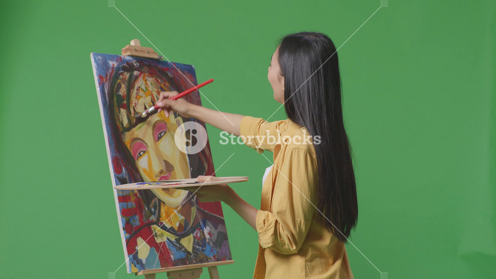 Asian female artist looking at the picture on canvas then saying wow while painting by oil paints and brush in the green screen studio