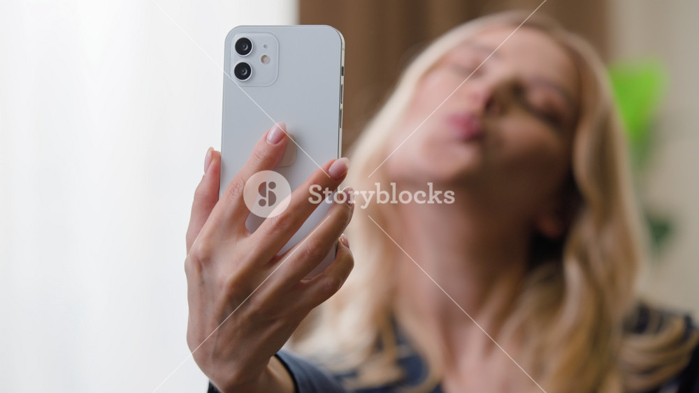 Happy pretty young Caucasian woman with mobile phone making selfie photo taking pictures photography kissing smiling grimacing for content