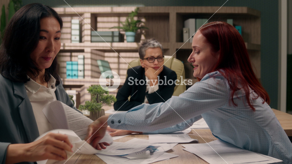 Calm Caucasian middle-aged woman senior boss looking at crazy multiethnic businesswomen throwing tearing business papers with stats chaotic