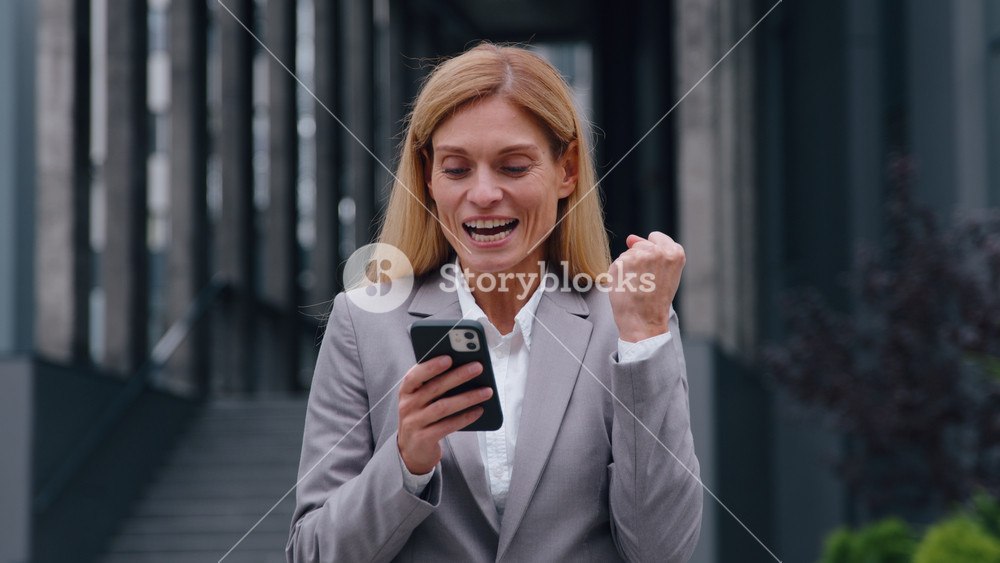 Surprised happy Caucasian adult 40s business woman with phone win in city amazed winner businesswoman winning with mobile app yes gesture