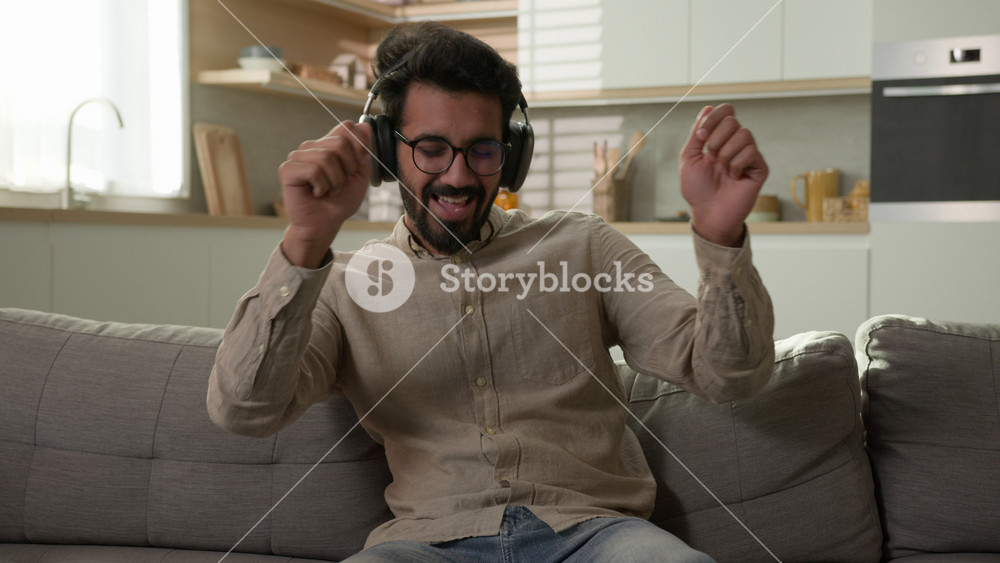 Happy relaxed Arabian man homeowner listen music in headphones enjoy rhythm song pretend playing drums on home sofa cheerful Indian guy