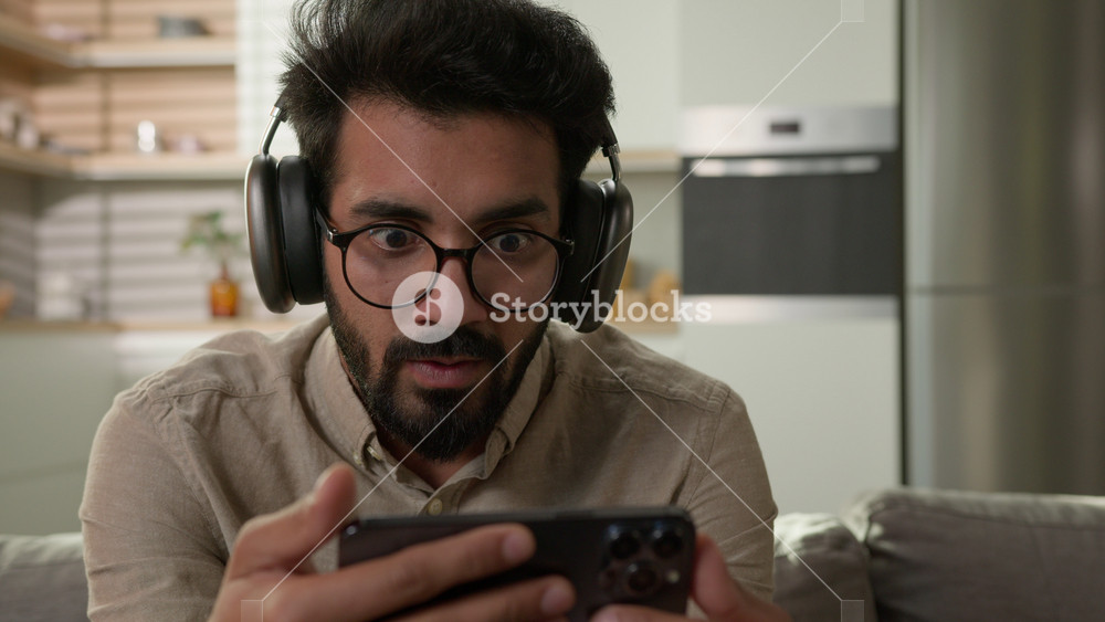 Surprised happy Arabian Indian man in headphones watch video stream on mobile phone excited male home owner watching football match online