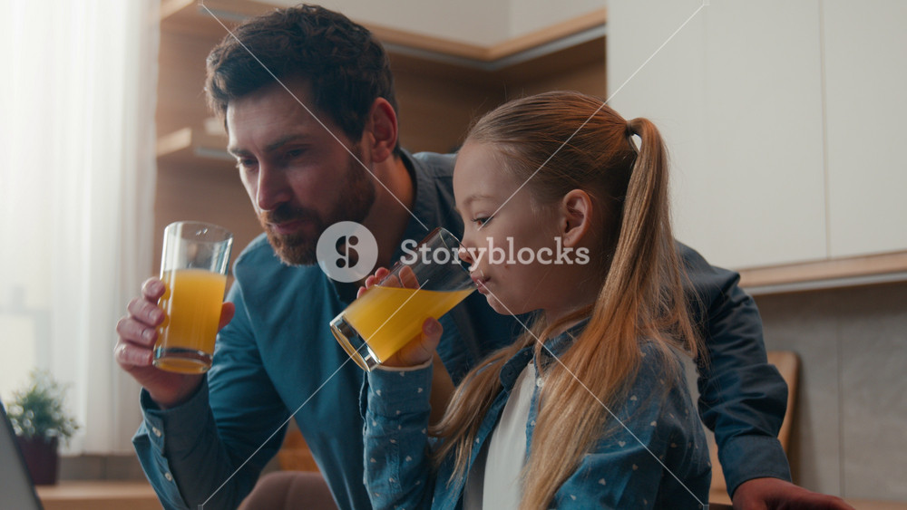 Caucasian happy family at kitchen adult father with little daughter child girl drinking fresh drink tasty orange juice looking at laptop dad