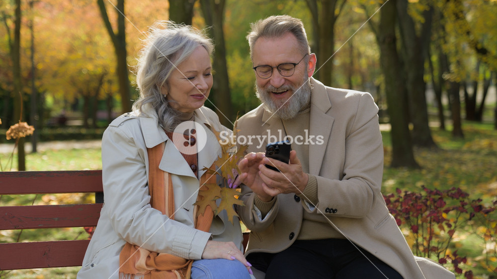 Caucasian retired couple family in love mature woman man sit on bench autumn park using mobile phone gadget together smiling wife husband