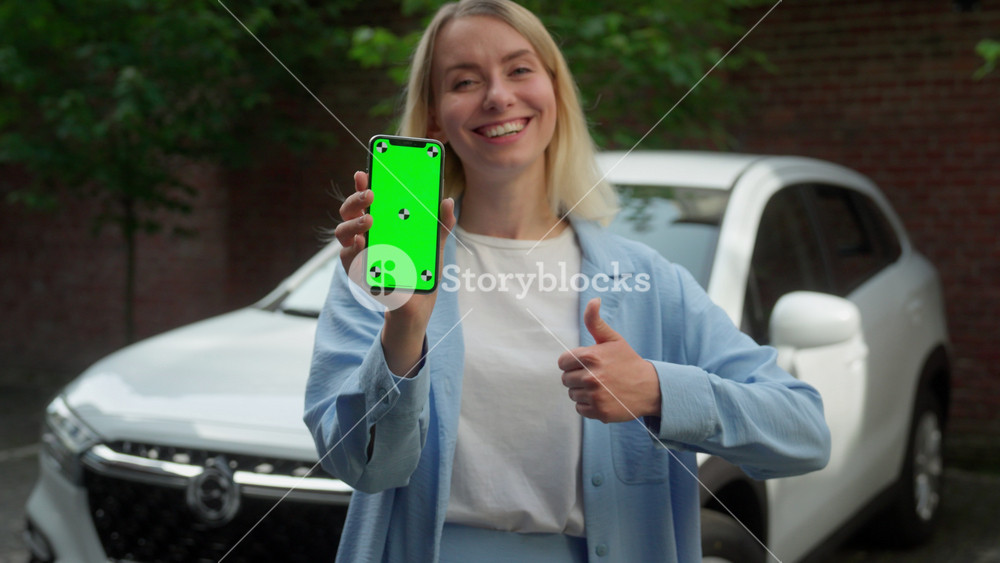 Smiling Caucasian girl female woman holding mobile phone tech green screen app chromakey automobile car driver showing like thumb up in city