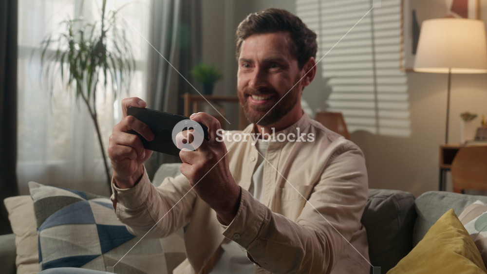Excited happy Caucasian adult man gamer playing mobile phone on couch sofa living room smiling guy male homeowner gaming addicted gadget