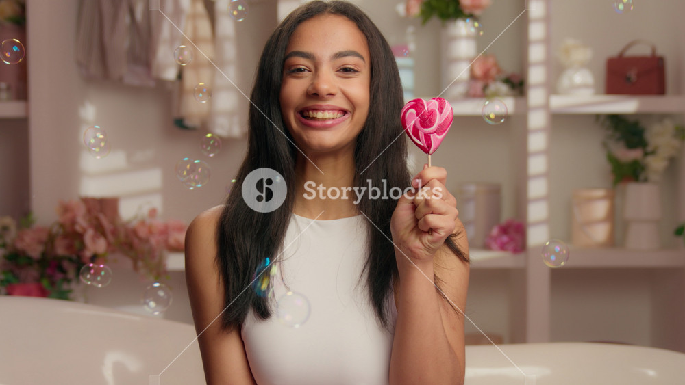 Portrait happy smiling American girl holding candy heart shape in pink bathroom flying soap bubbles close up positive Hispanic young woman