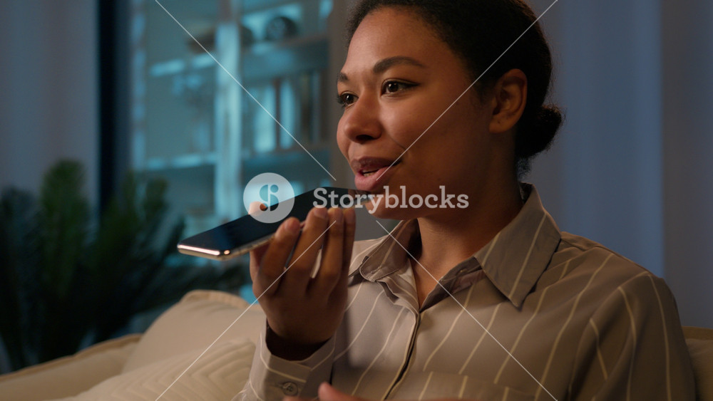African American woman happy smiling ethnic girl record voice message on speaker phone dictate audio on smartphone at evening night home say