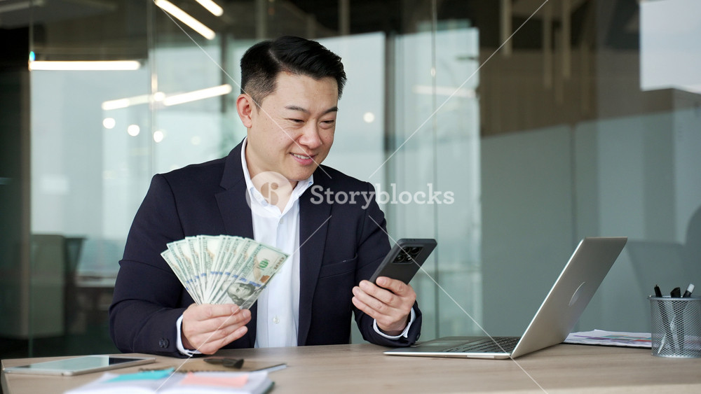 Happy asian businessman holding fan of cash money dollar bills and mobile phone sitting in a business office looking at camera. Male is satisfied with earning money using application on his smartphone