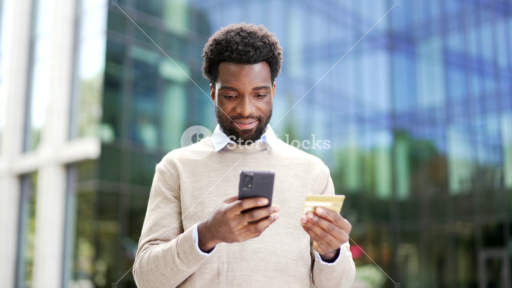 Smiling african american businessman is shopping online using credit card and mobile phone standing on street near business office building. Worker happy about successful transaction or good price