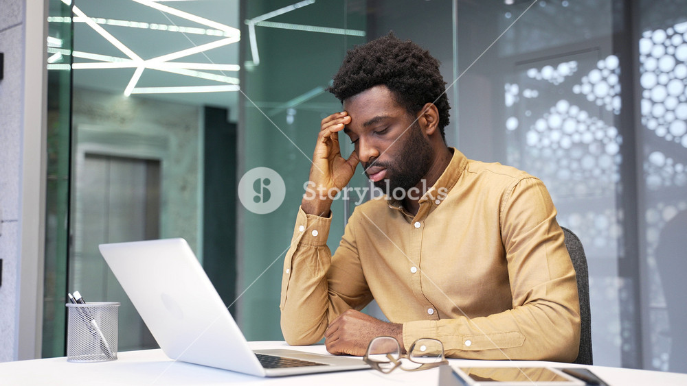 Depressed African American businessman feels anxiety and mental pain at work. Lonely manager sitting at corporate office desk experiences severe emotional distress and burnout indoors.