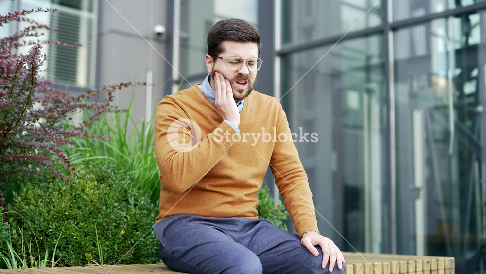 Businessman suffering from toothache sitting on bench on street near business office building. Upset adult bearded man holds his hand on his cheek, massaging sore spot, needs treatment at the dentist