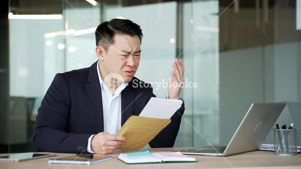 Worried asian businessman reading letter with bad news sitting at desk at workplace in business office. Disappointed upset entrepreneur in formal suit frustrated by received negative notification
