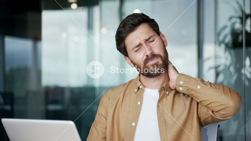 Tired businessman suffering from neck pain while sitting at desk at workplace in business office. Upset male employee feels discomfort, massages and rubs sore muscles, stretches himself Close up