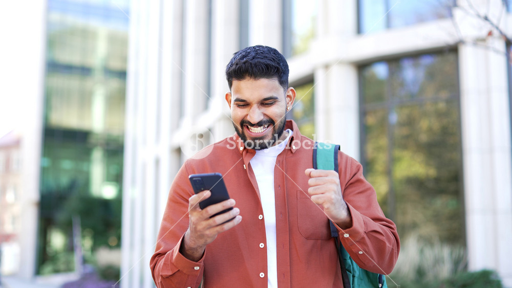 Satisfied happy male student received great news on mobile phone standing outside in the campus space near university building. Smiling joyful handsome man reads positive good message on smartphone