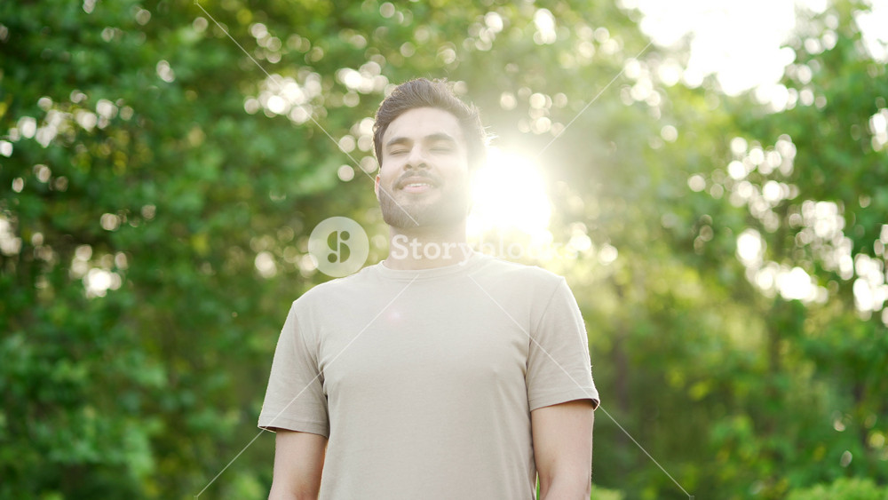 Happy young adult sportsman relaxing with closed eyes standing in urban city park. Smiling handsome fit man breathes deeply, feels relief and enjoys being in nature while resting after morning workout