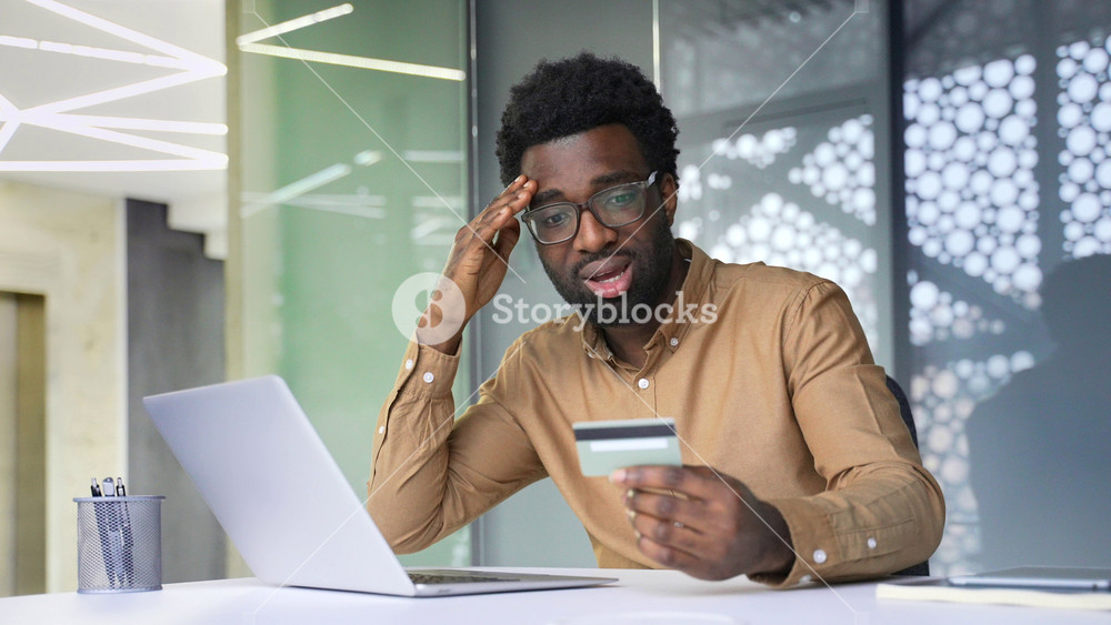 Shocked African American businessman holds credit card and looks at laptop screen. Frustrated black man manager faces financial fraud, online scam and stolen money from bank account indoors.