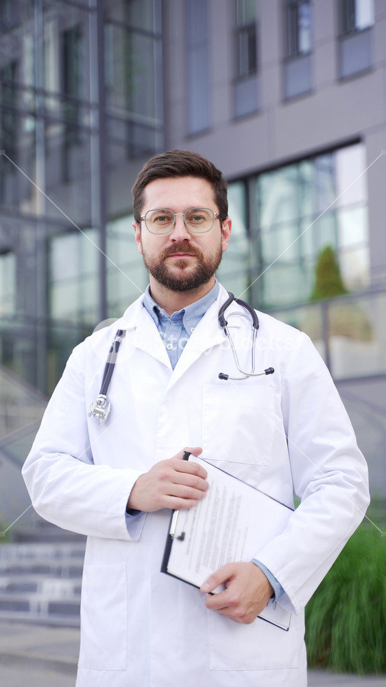 Portrait of a serious doctor in a white coat looking at camera, standing outside near the hospital building. Confident male medical worker physician posing on street holding a folder. Vertical video