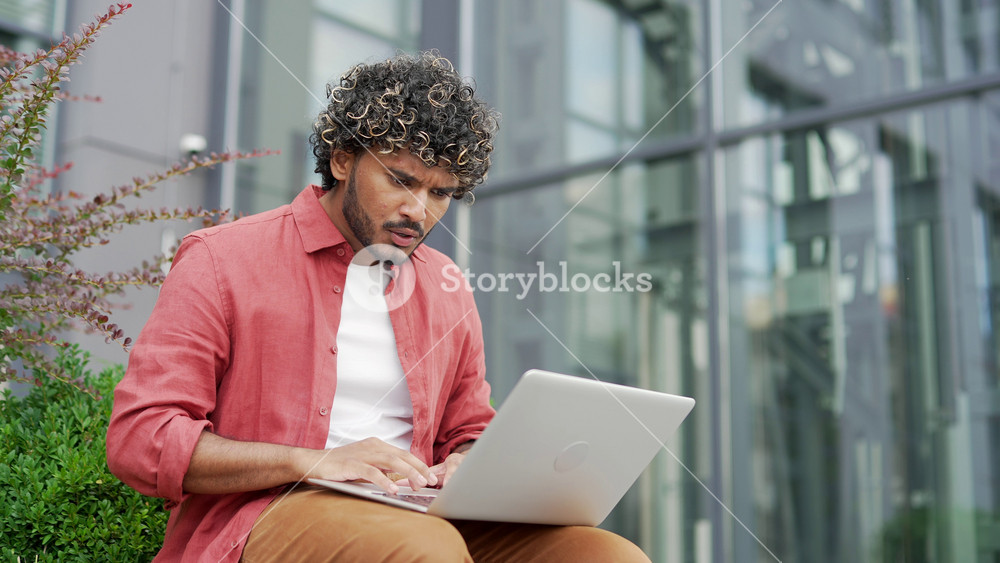 Shocked worried young businessman reading bad news on laptop sitting on a bench on the street near modern office building. Frustrated stressed handsome man upset looking at negative message. Close up