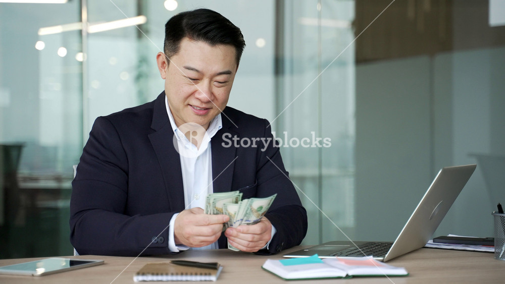 Smiling asian businessman in a formal suit counting dollar bills while sitting at a desk at a workplace inbusiness office. Happy satisfied worker holding cash in hands, calculates his earnings, salary